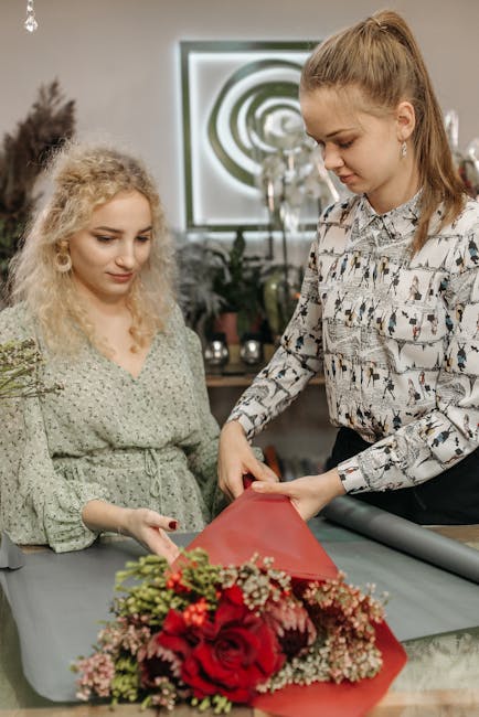 Two women are engaged in a floral arrangement in an indoor setting, with a large bouquet of vibrant red roses, pink and white filler flowers, and green foliage wrapped in red tissue paper. The woman o