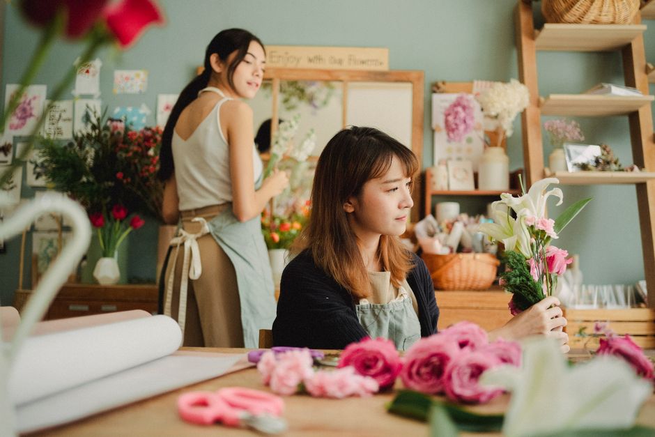 A florist workshop scene at flowershook.co.uk showing two women arranging floral bouquets inside a well-lit shop. The woman in the foreground, with shoulder-length brown hair and wearing a dark cardig