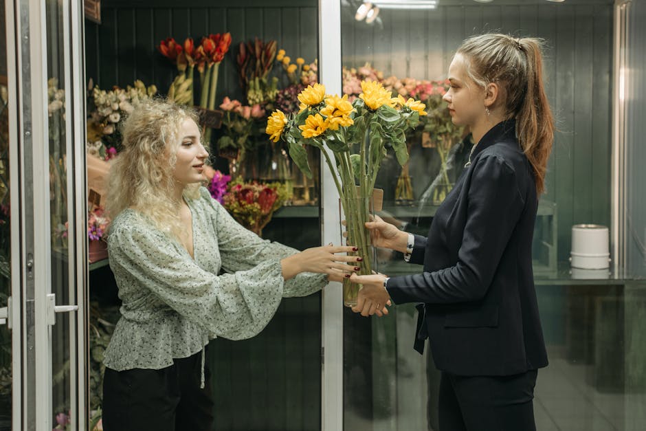 A florist in a dark blazer is handing over a tall bouquet of bright yellow sunflower flowers to a woman with curly blonde hair wearing a light-colored, floral-patterned blouse inside a flower shop. Th