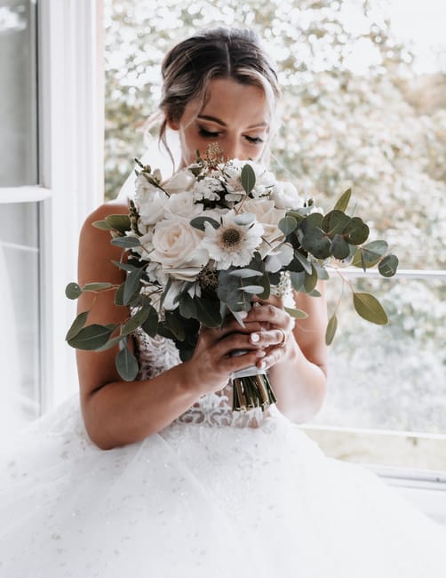 A bride holding a large, elegant bouquet featuring predominantly white flowers, including roses and anemones, accented with soft green eucalyptus leaves. The bouquet is wrapped with a simple ribbon, a