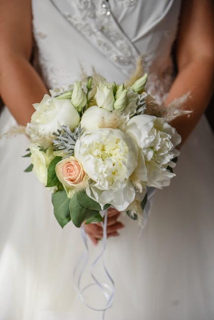 A bridal bouquet consisting of white and blush pink flowers, including roses and peonies, with delicate green foliage and soft grey accents. The arrangement is held by a bride dressed in a silver-embe