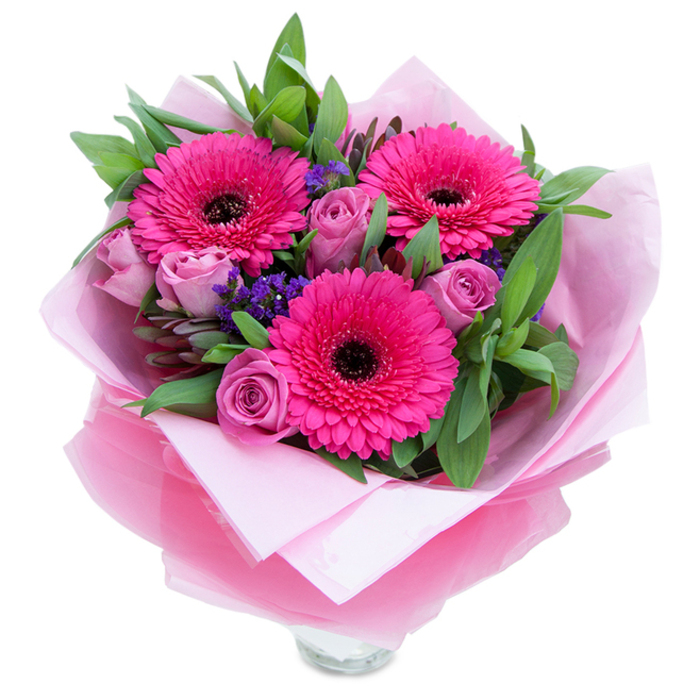 I often think of bouquets as gestures that travel: this one would arrive in Hook like a warm note. At its centre are three assertive Gerbera daisies in a saturated fuchsia - petals layered like small fans around dark, near-black centres that punctuate the colour with dramatic contrast. Clustered around them, roses in a soft lavender-pink or dusty-rose hue open in controlled spirals, their velvety petals creating gentle shadows and a tactile softness you want to touch. Little clusters of purple statice are sprinkled throughout, their papery texture and cooler tones adding depth and the suggestion of wildflower charm. The greenery behind and between the blooms is generous and leafy, with broad salal-style leaves and the subtle grey-green of eucalyptus that lend a slightly aromatic, dewy note. The arrangement is wrapped for gifting: an inner layer of matte pale-pink tissue cradles the stems while a translucent, slightly crinkled fuchsia-pink cellophane wraps around, catching the light and tying the composition together visually. The lower edge reveals the top rim of a clear glass vase, hinting at immediate display - perhaps on a kitchen island near Hook village green or on a sideboard along Basingstoke Road. The overall impression is playful yet graceful; the textures and scents combine to create a bright, optimistic bouquet suited to birthdays, anniversaries, or a spontaneous 'you're special' delivery. As someone who arranges flowers for local homes and small events in the Hart area, I see this as a dependable, mood-lifting selection that reads as thoughtful and well-made.